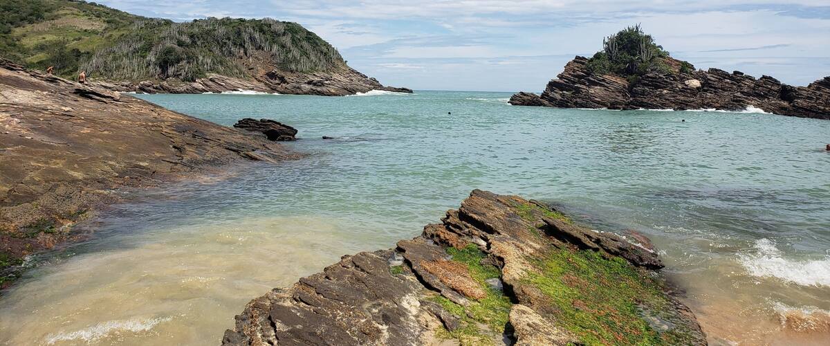 A coastal landscape with rocky shores, clear turquoise waters, and a hilly backdrop under a partly cloudy sky.