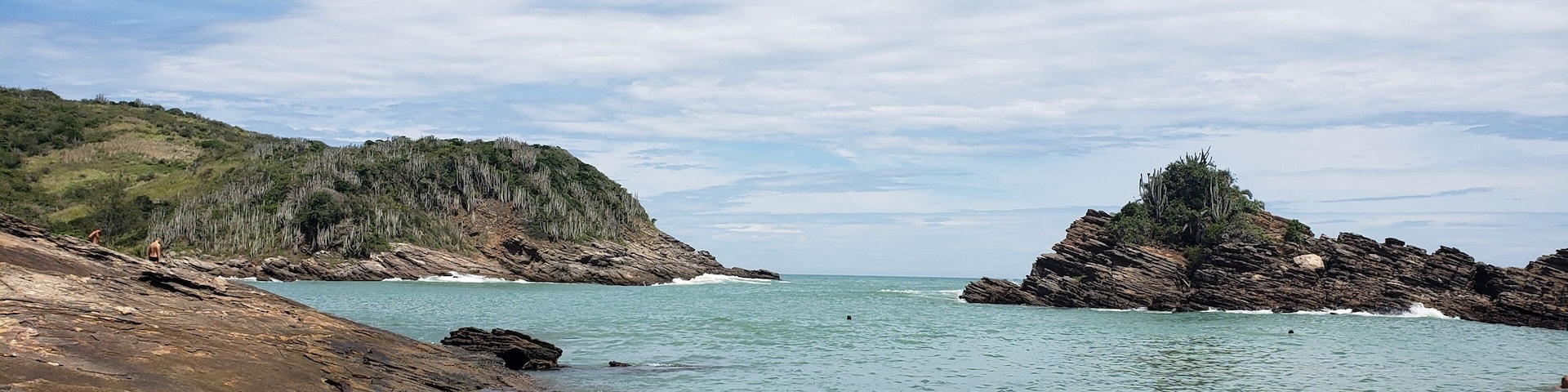 A coastal landscape with rocky shores, clear turquoise waters, and a hilly backdrop under a partly cloudy sky.
