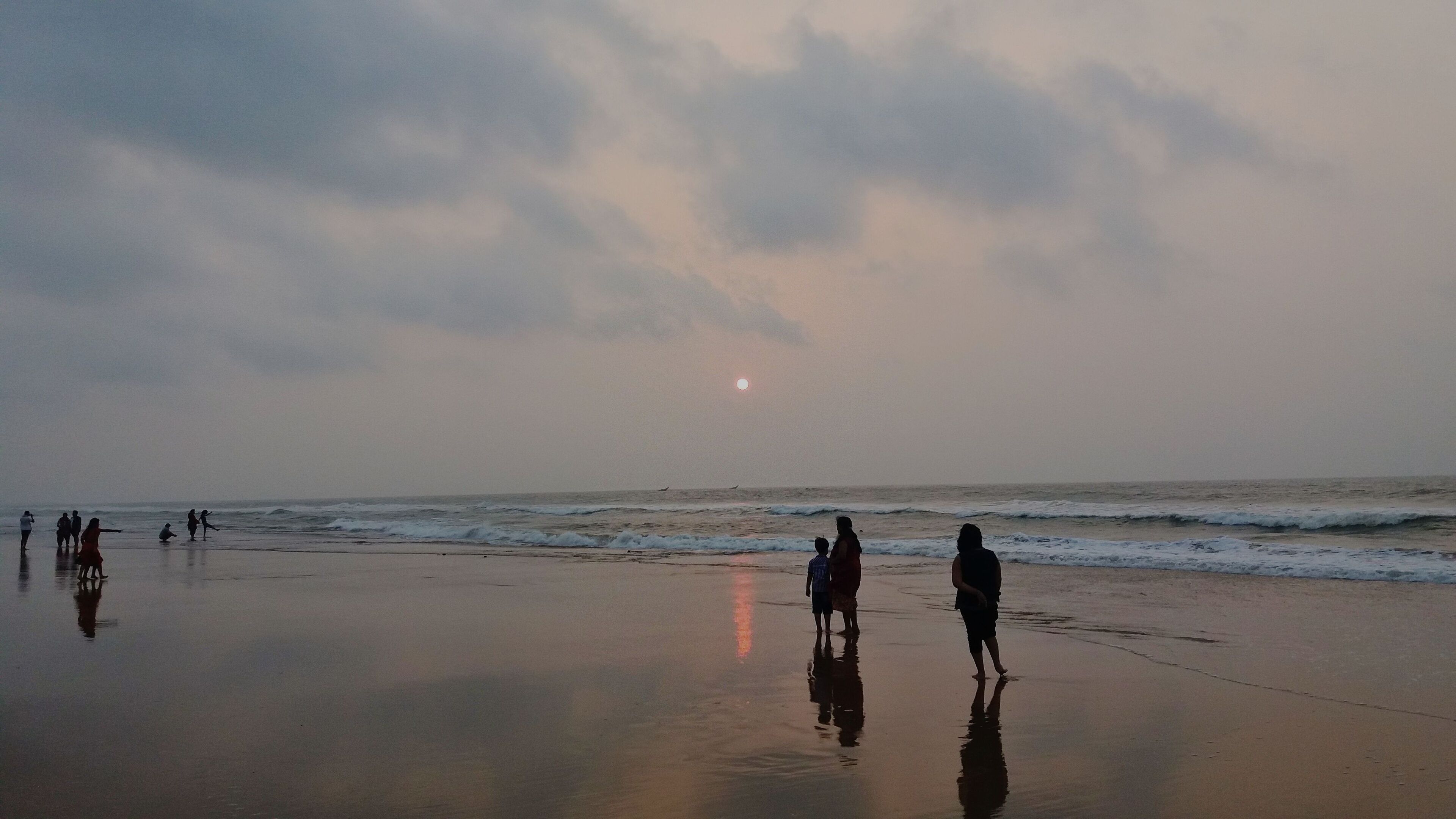 A beach with people walking and playing in the sand, waves crashing onto the shore, and a sunset in the background.