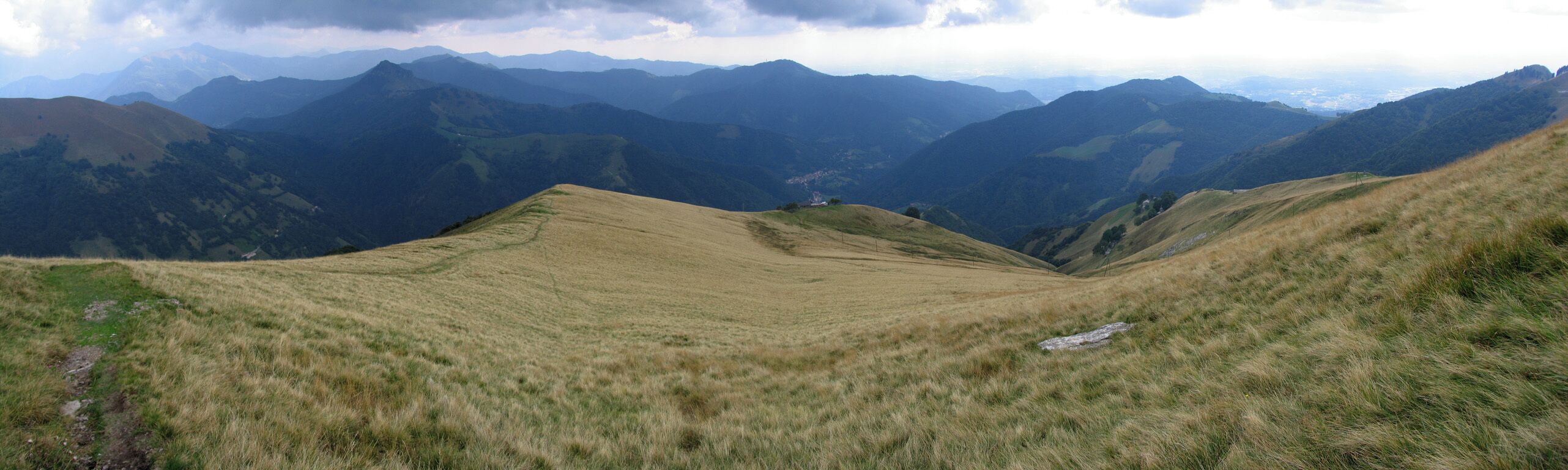 A grassy valley with rolling hills and distant mountain ranges under a cloudy sky.