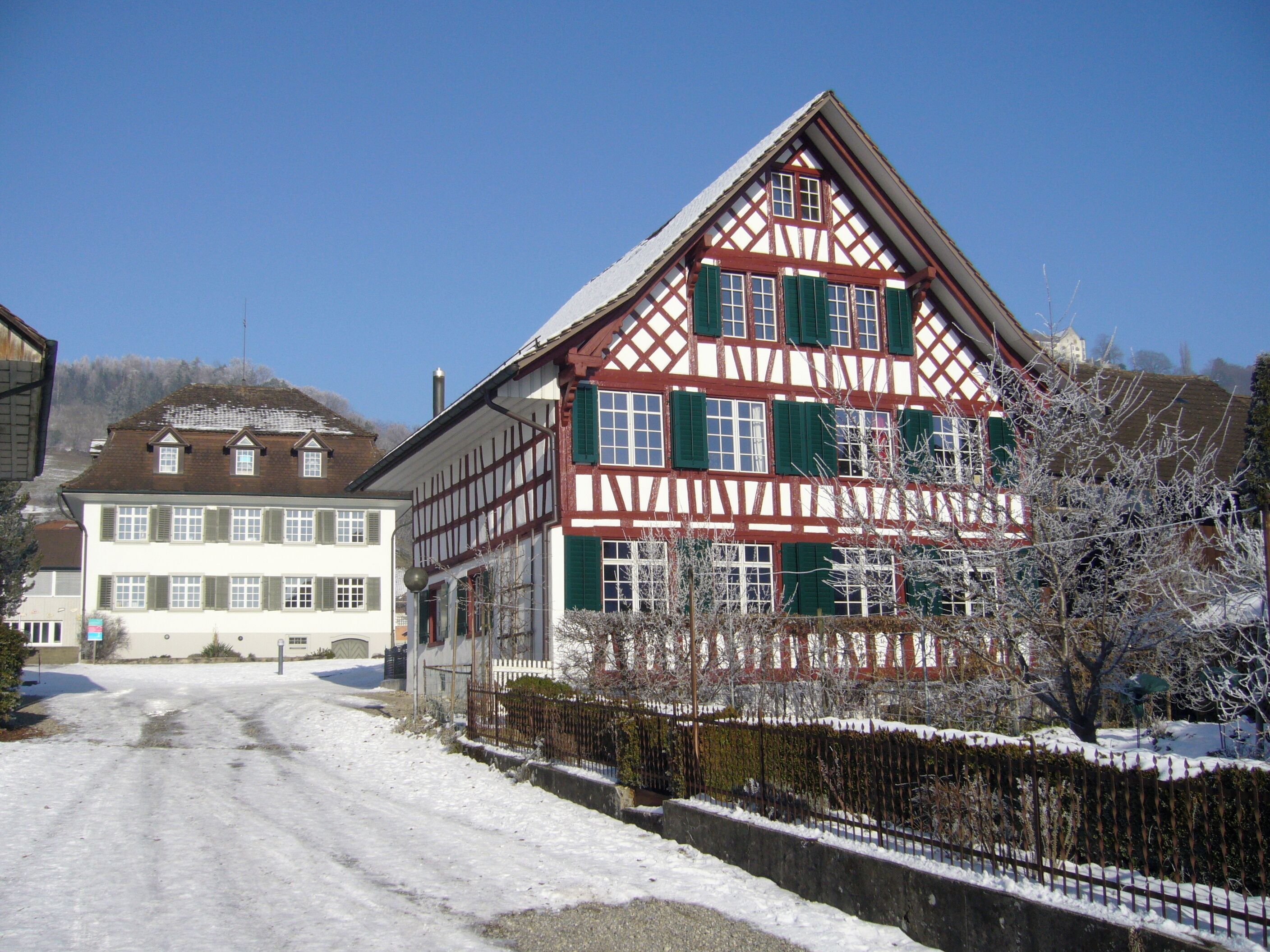 A traditional half-timbered house with green shutters, a snow-covered driveway, and a clear blue sky.