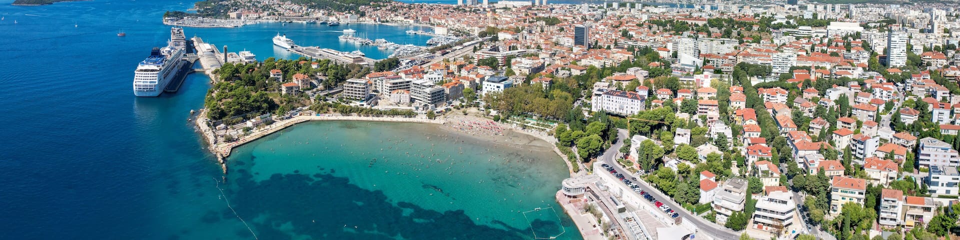 A coastal city with a large cruise ship docked, clear blue waters, and a mountainous backdrop.