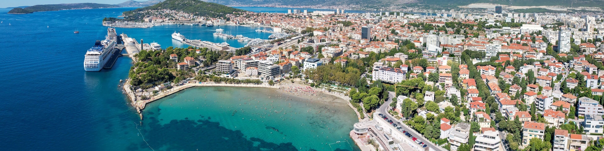 A coastal city with a large cruise ship docked, clear blue waters, and a mountainous backdrop.