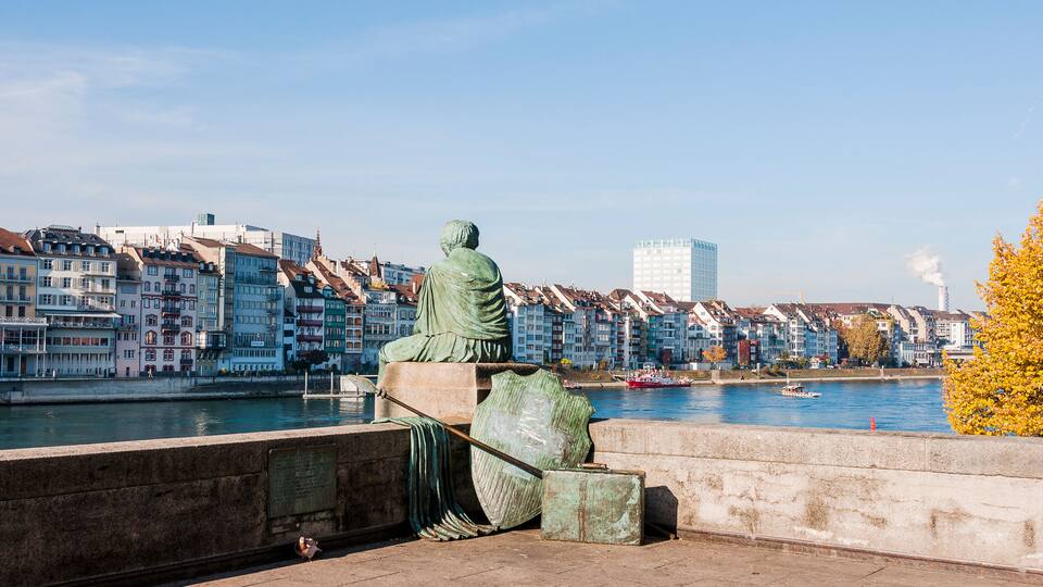 Estátua de uma figura sentada com vista para um rio e edifícios ao fundo.