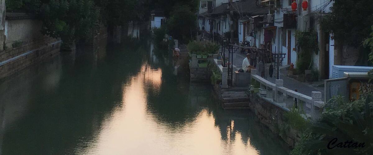 A calm river at sunset with traditional buildings on the right and dense foliage on the left.
