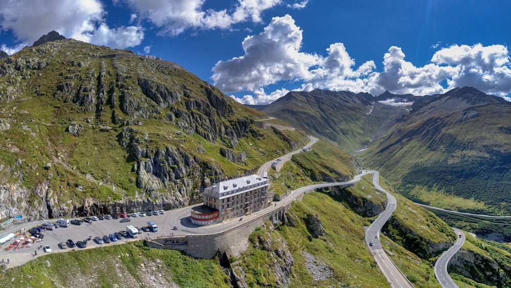 A mountain road winding through a valley with a parking area and a building on the left.