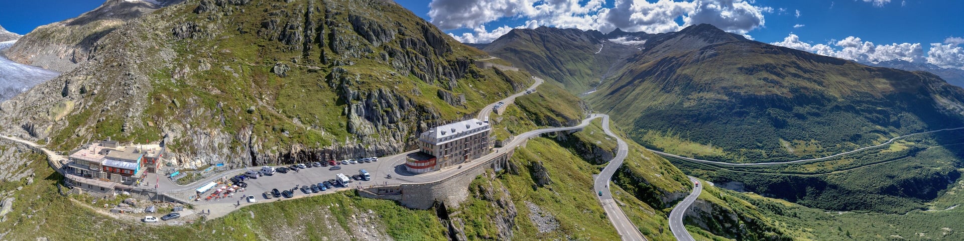 A mountain road winding through a valley with a parking area and a building on the left.