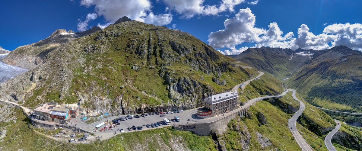 A mountain road winding through a valley with a parking area and a building on the left.