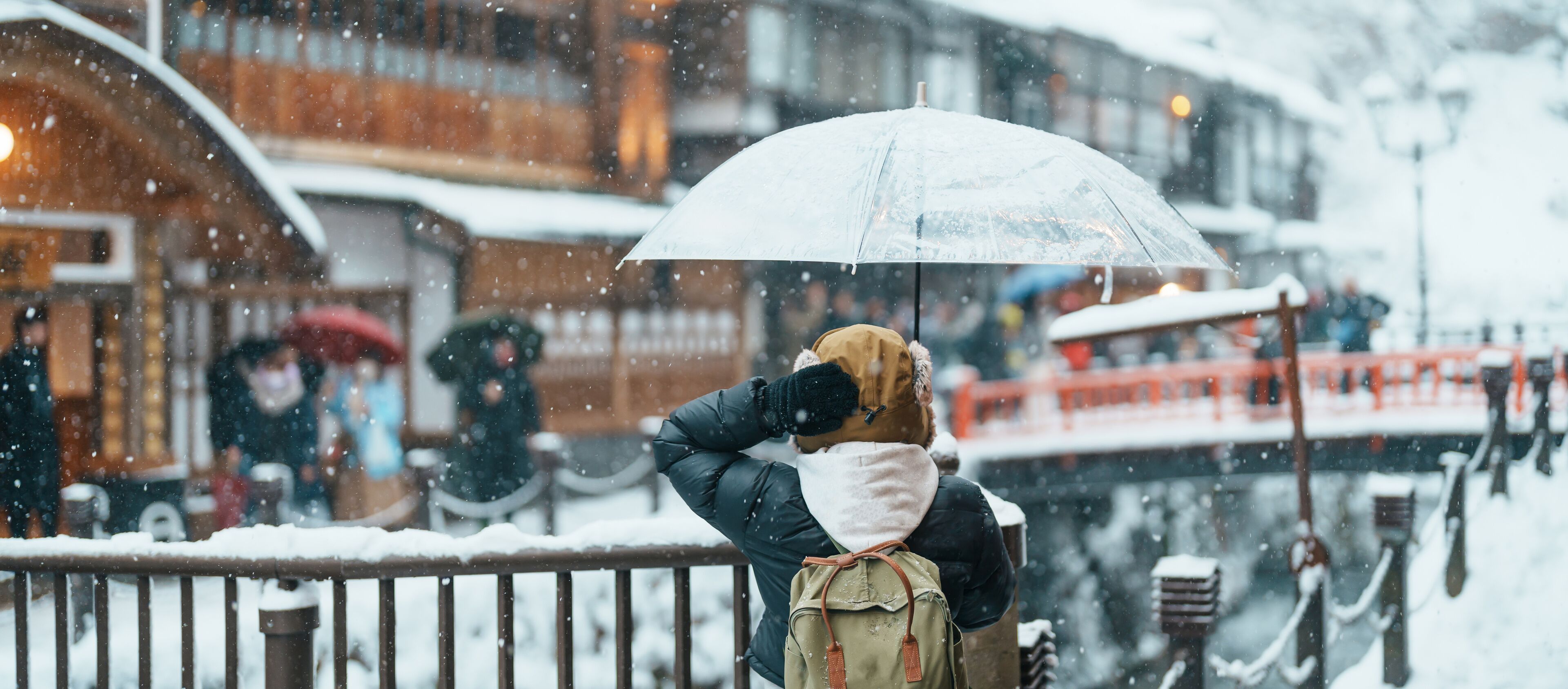 A person standing on a snow-covered bridge, holding a transparent umbrella.
