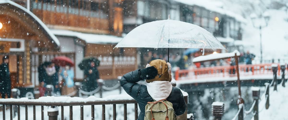 A person standing on a snow-covered bridge, holding a transparent umbrella.