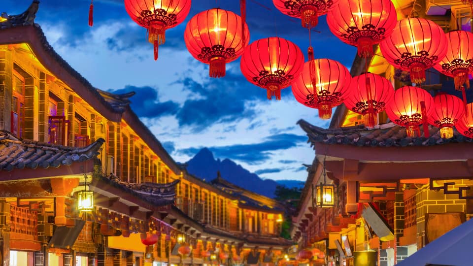 A traditional Chinese street at dusk with red lanterns, ornate rooftops, and a mountain backdrop.