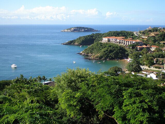 A coastal landscape with a large building, lush greenery, and calm sea.