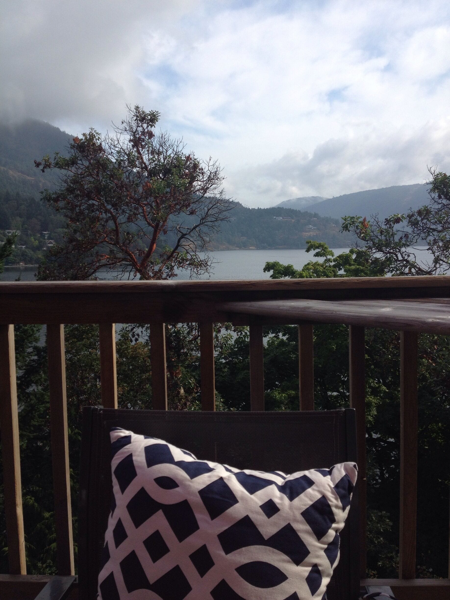 A wooden deck with a patterned pillow, overlooking a lake with mountains in the background.