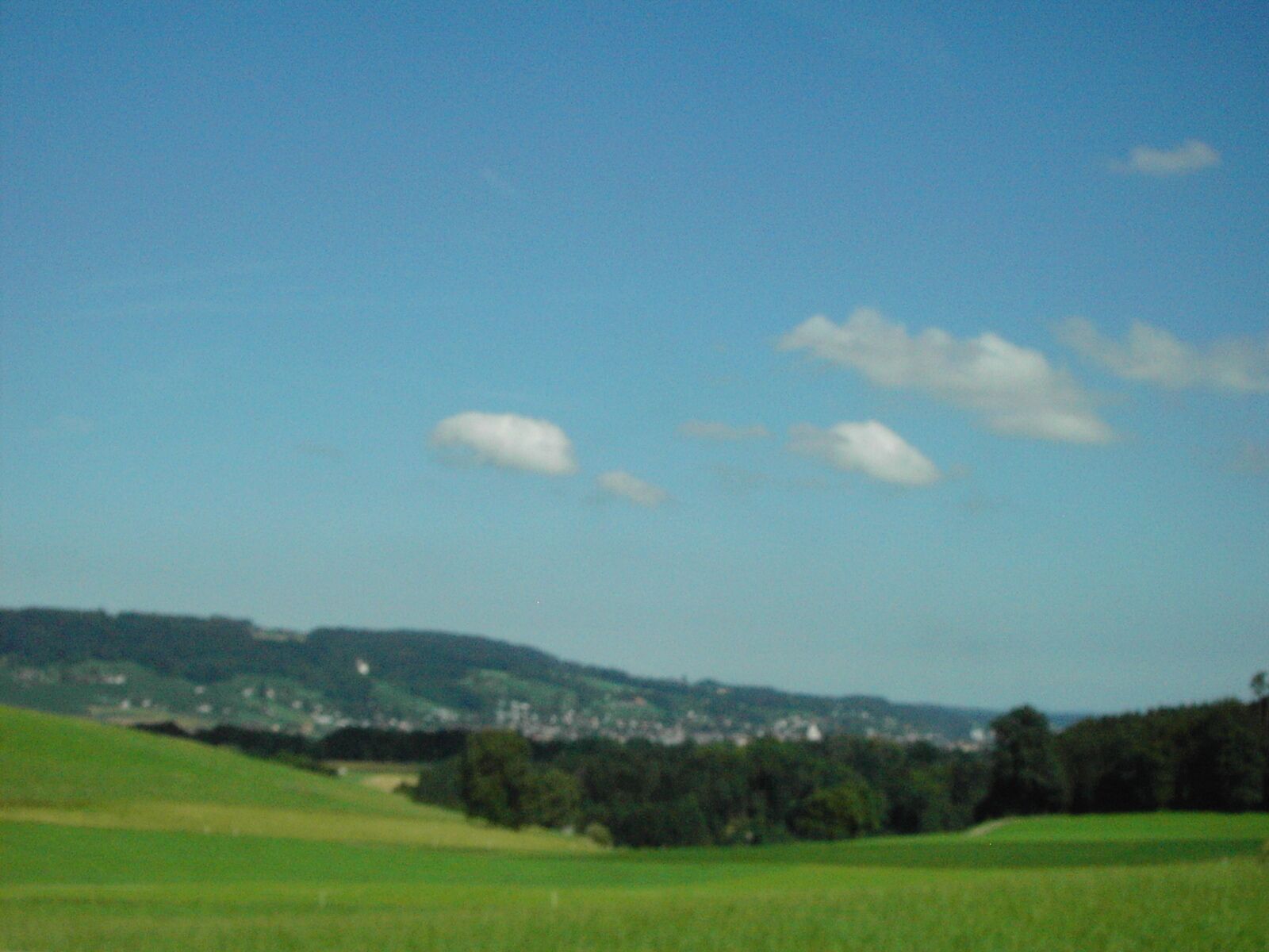 A green field with a town in the distance and a hill with trees.