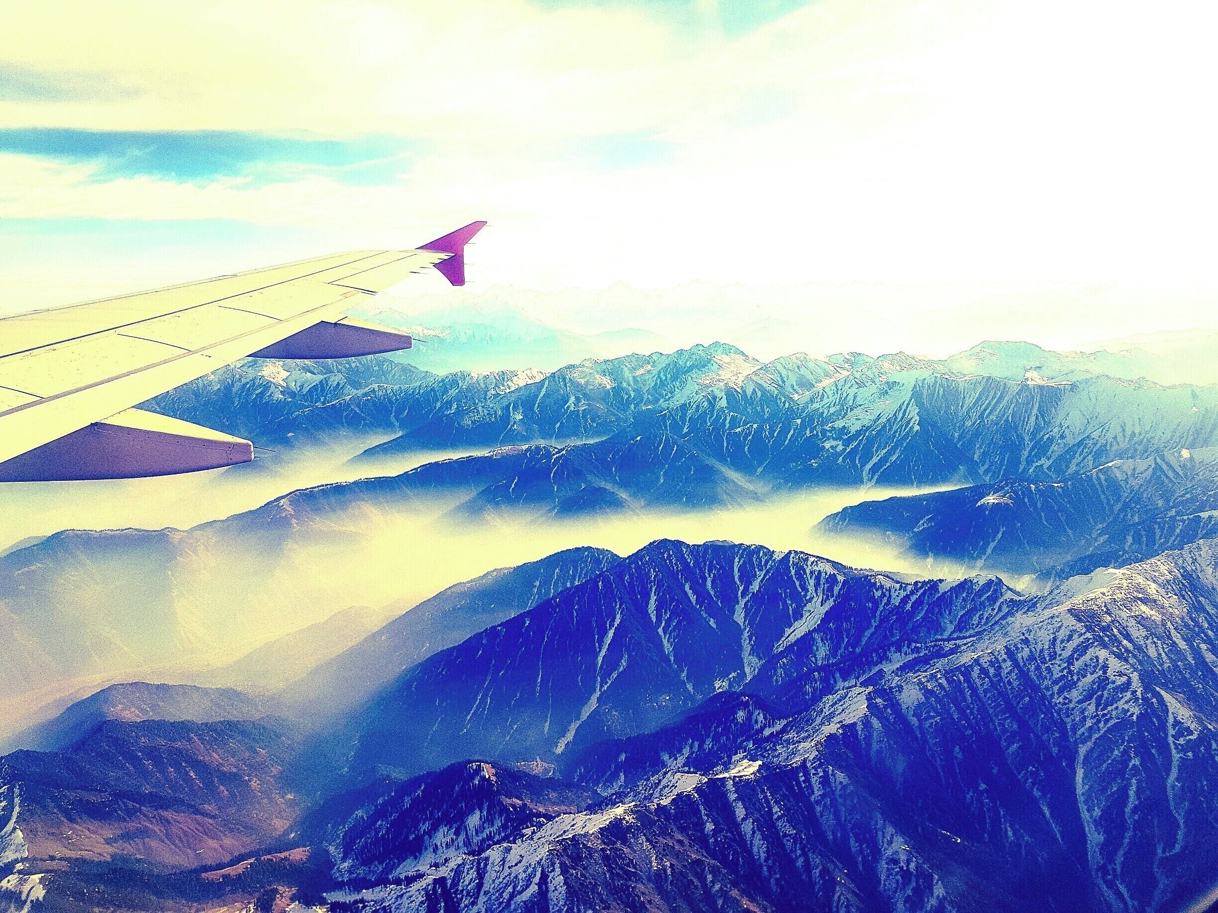 An airplane wing with a purple tip flying over snow-covered mountains and valleys.