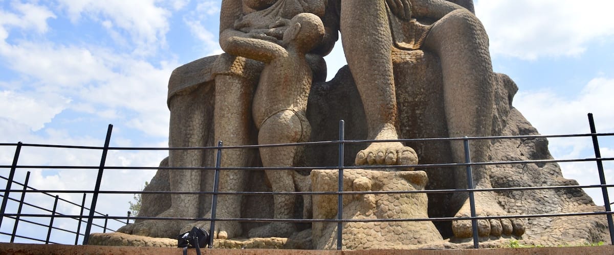 A large stone sculpture of two figures seated on a rock, surrounded by a metal fence.