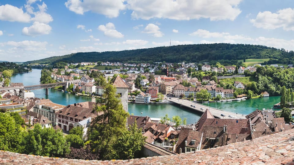 A riverside town with historic buildings, a bridge, and a hill in the background.