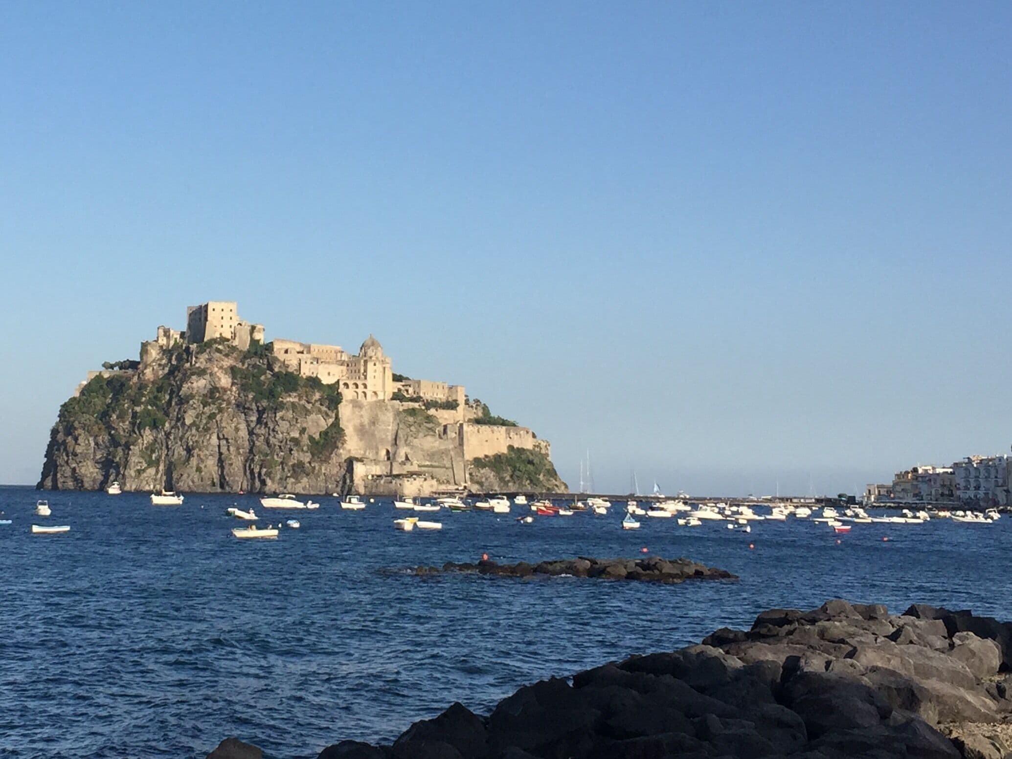 Un pueblo costero con un castillo imponente en una isla rocosa, rodeado de barcos y un cielo azul despejado.