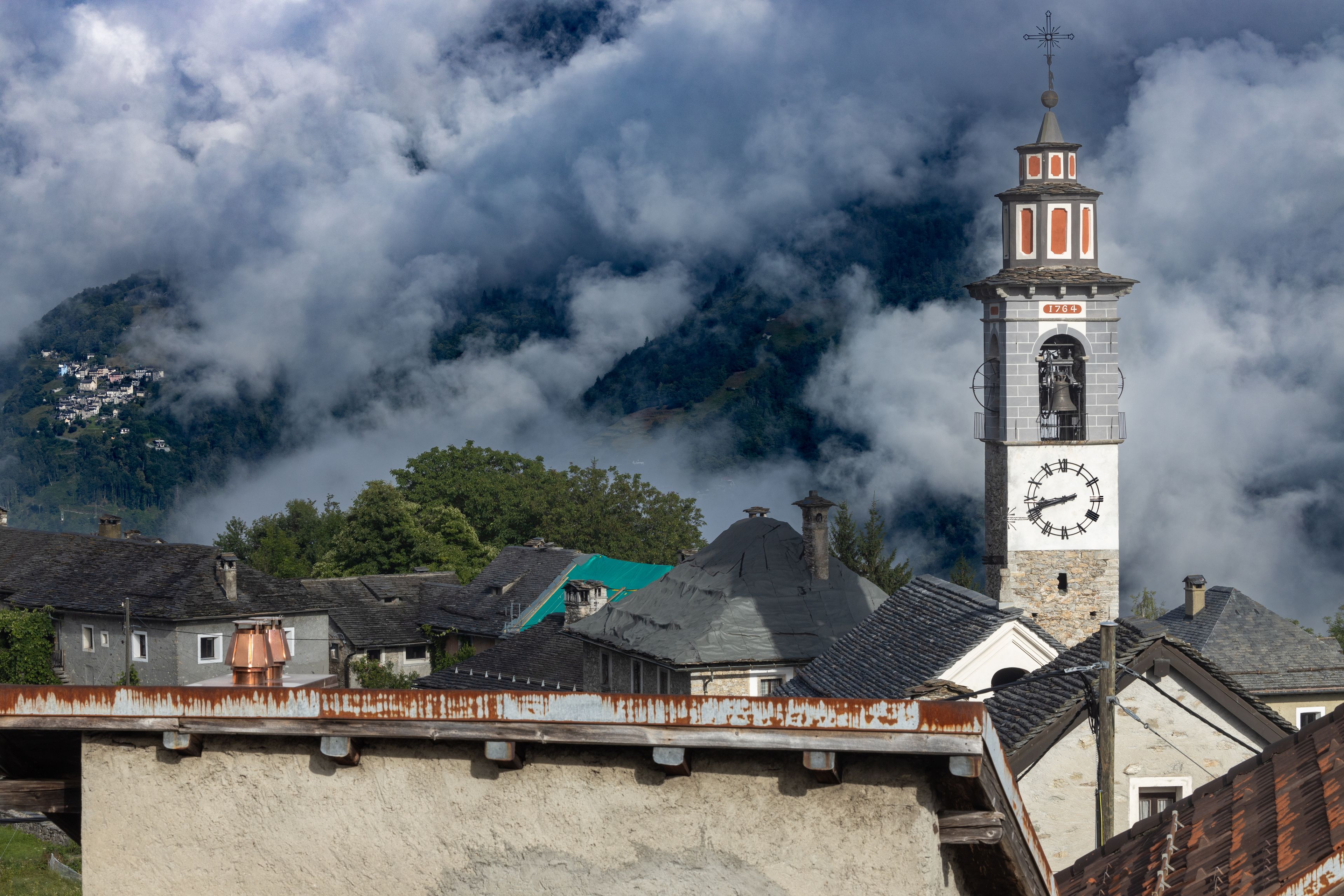 Ein Glockenturm mit Wetterfahne, umgeben von einem Dorf mit traditioneller Architektur und einem Berg im Hintergrund.