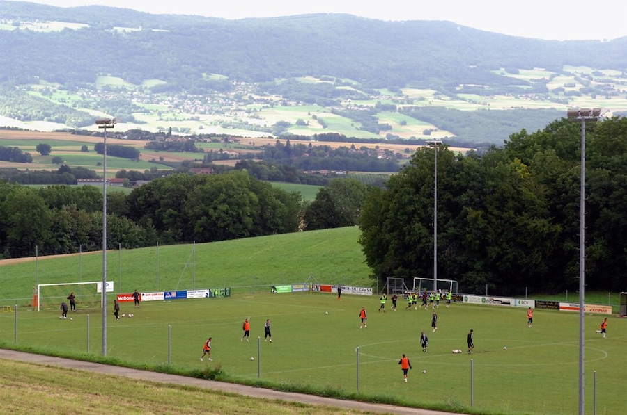 A soccer field with players, surrounded by trees and a hilly landscape in the background.