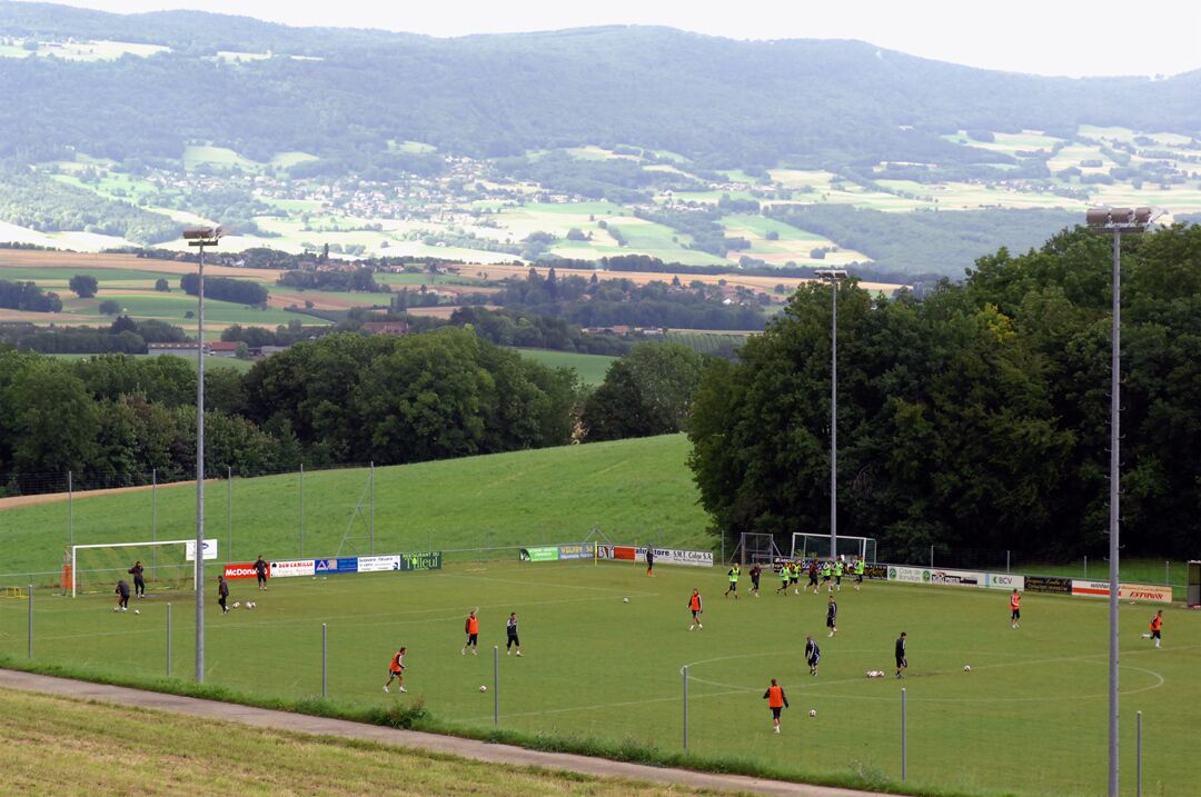 A soccer field with players, surrounded by trees and a hilly landscape in the background.