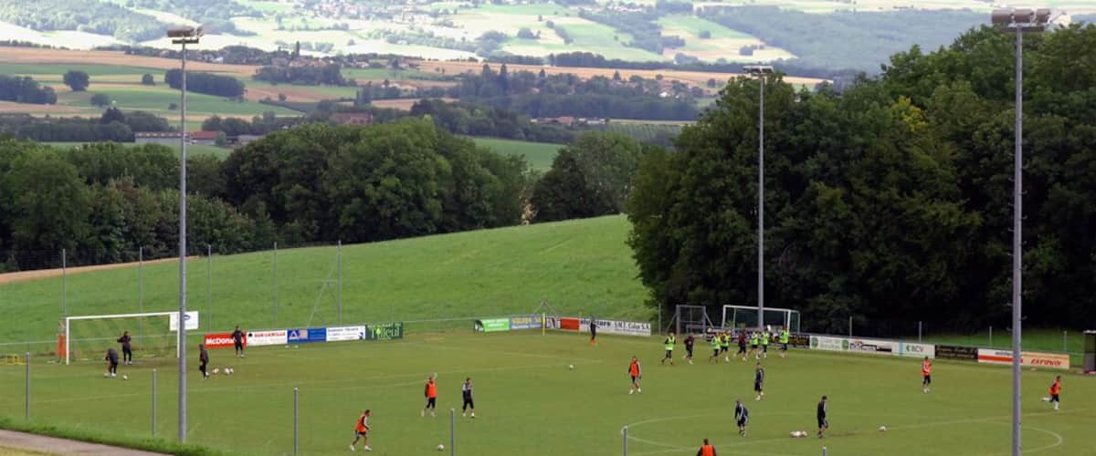 A soccer field with players, surrounded by trees and a hilly landscape in the background.
