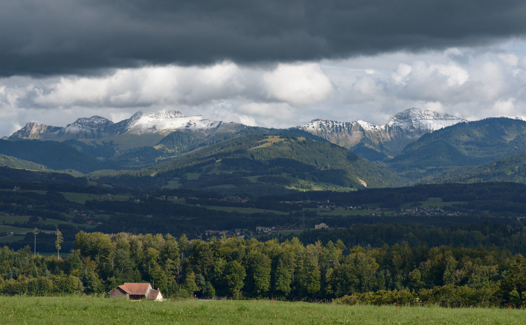 A mountainous landscape with snow-capped peaks, a forested valley, and a small building in the foreground.