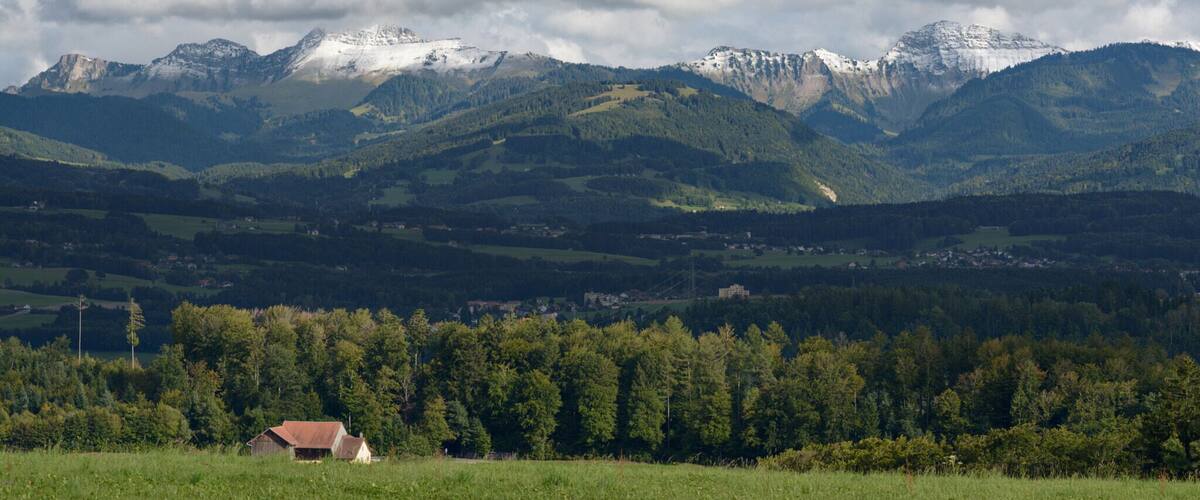 A mountainous landscape with snow-capped peaks, a forested valley, and a small building in the foreground.