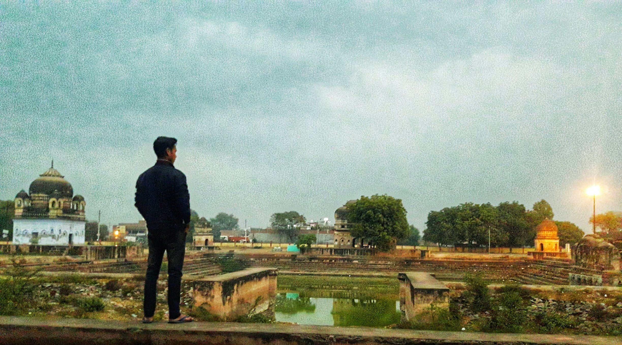 A man standing on a bridge overlooking a cityscape with domed buildings and a large open area with a pond.
