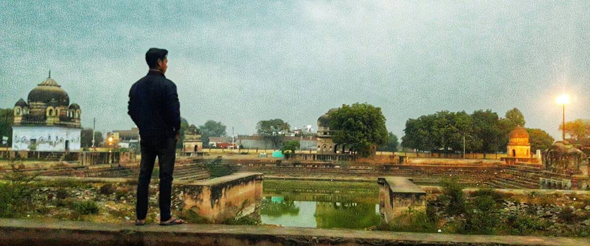 A man standing on a bridge overlooking a cityscape with domed buildings and a large open area with a pond.