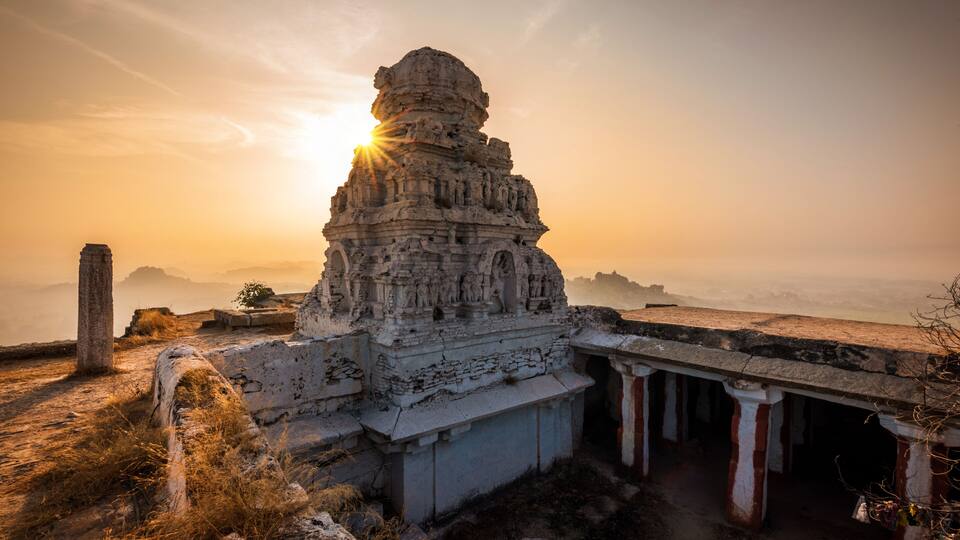 A temple with intricate carvings, a stone pillar, and a sunrise in the background.