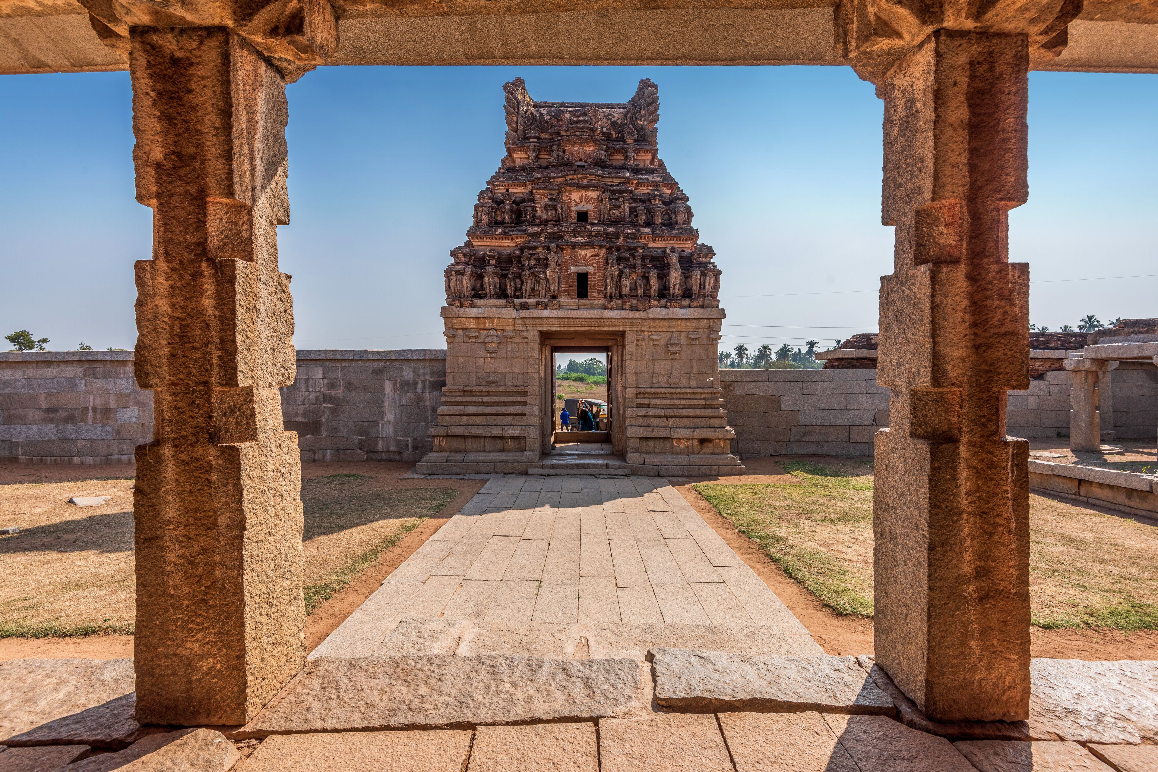 An ancient stone structure with intricate carvings, a paved pathway leading to it, and a clear blue sky in the background.