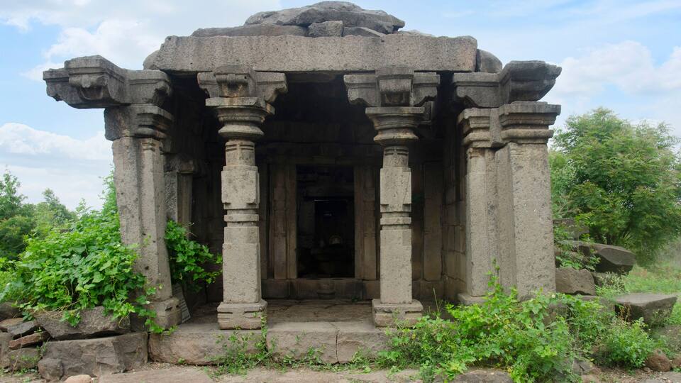 An ancient stone structure with a central doorway, flanked by columns, surrounded by greenery and rocks.