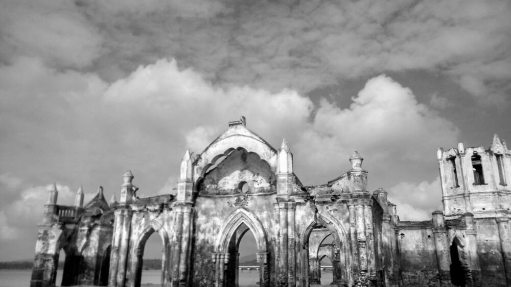 An abandoned, dilapidated building with Gothic architecture, including pointed arches and a central dome, surrounded by water.