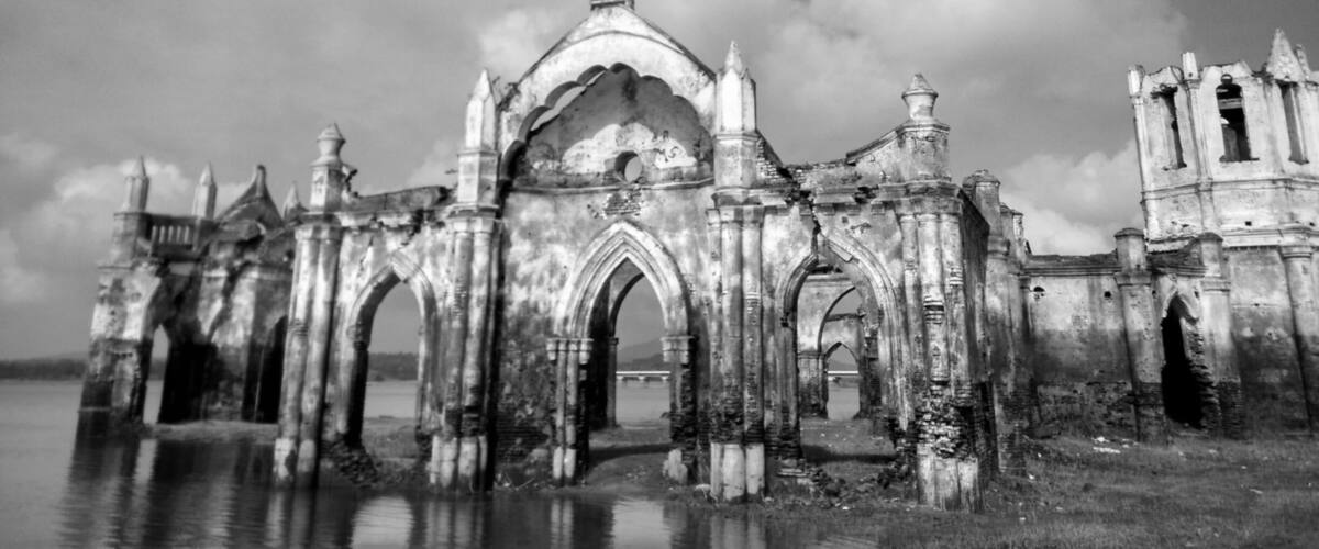 An abandoned, dilapidated building with Gothic architecture, including pointed arches and a central dome, surrounded by water.