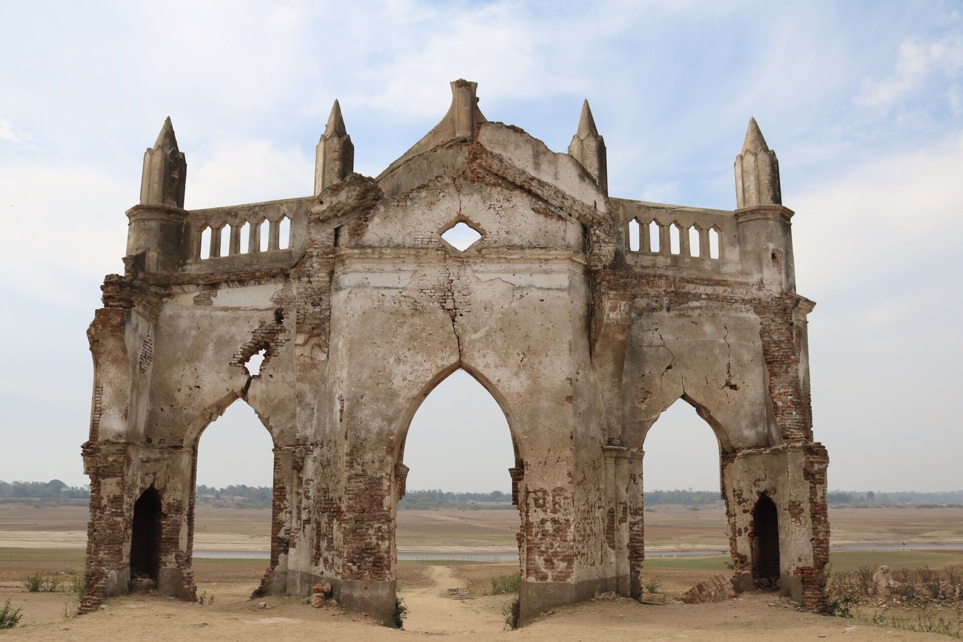 An ancient, weathered stone structure with multiple arches and pointed towers, set against a flat landscape.