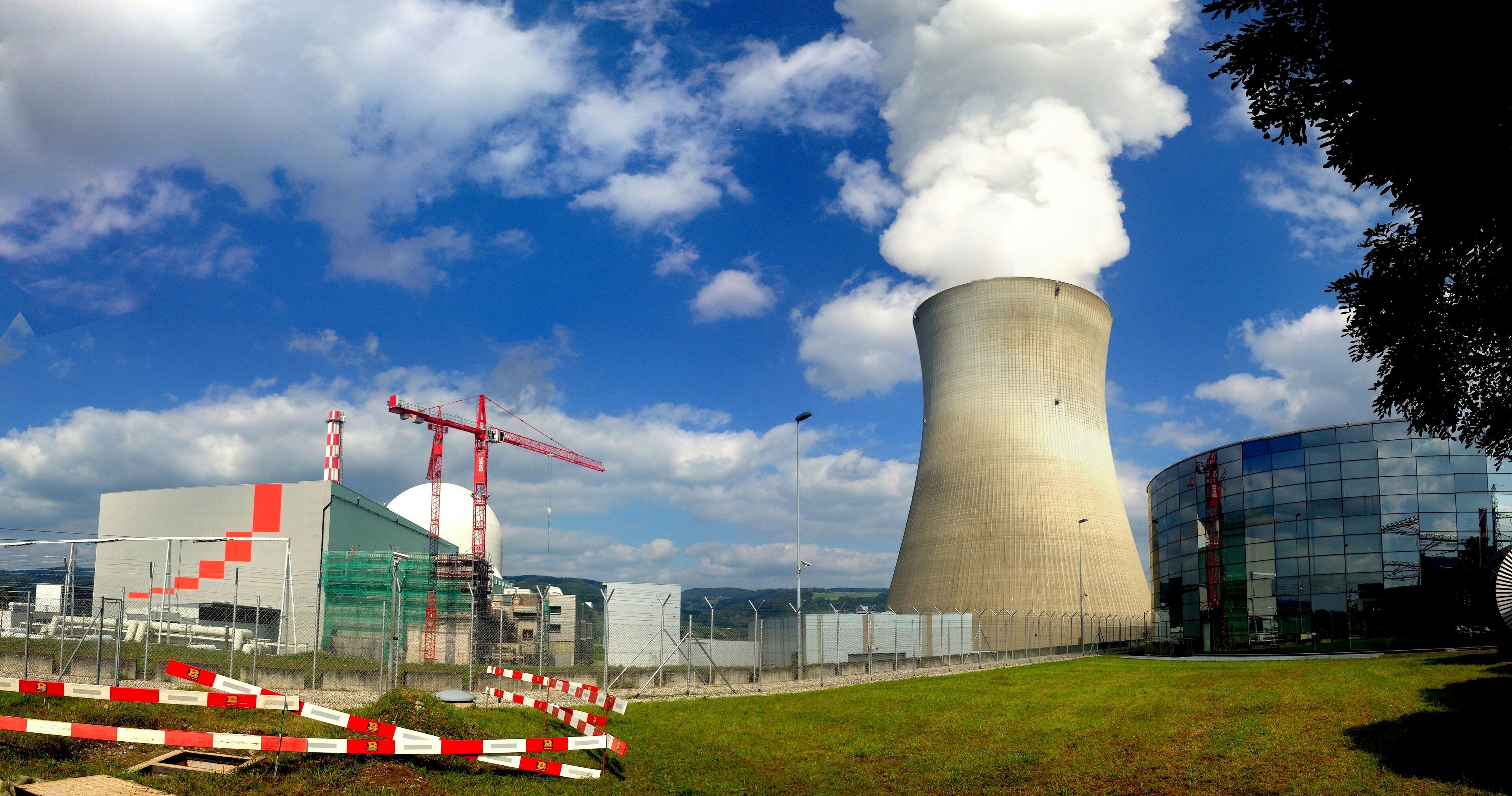 A nuclear power plant with cooling towers emitting steam, surrounded by construction barriers and a modern building.