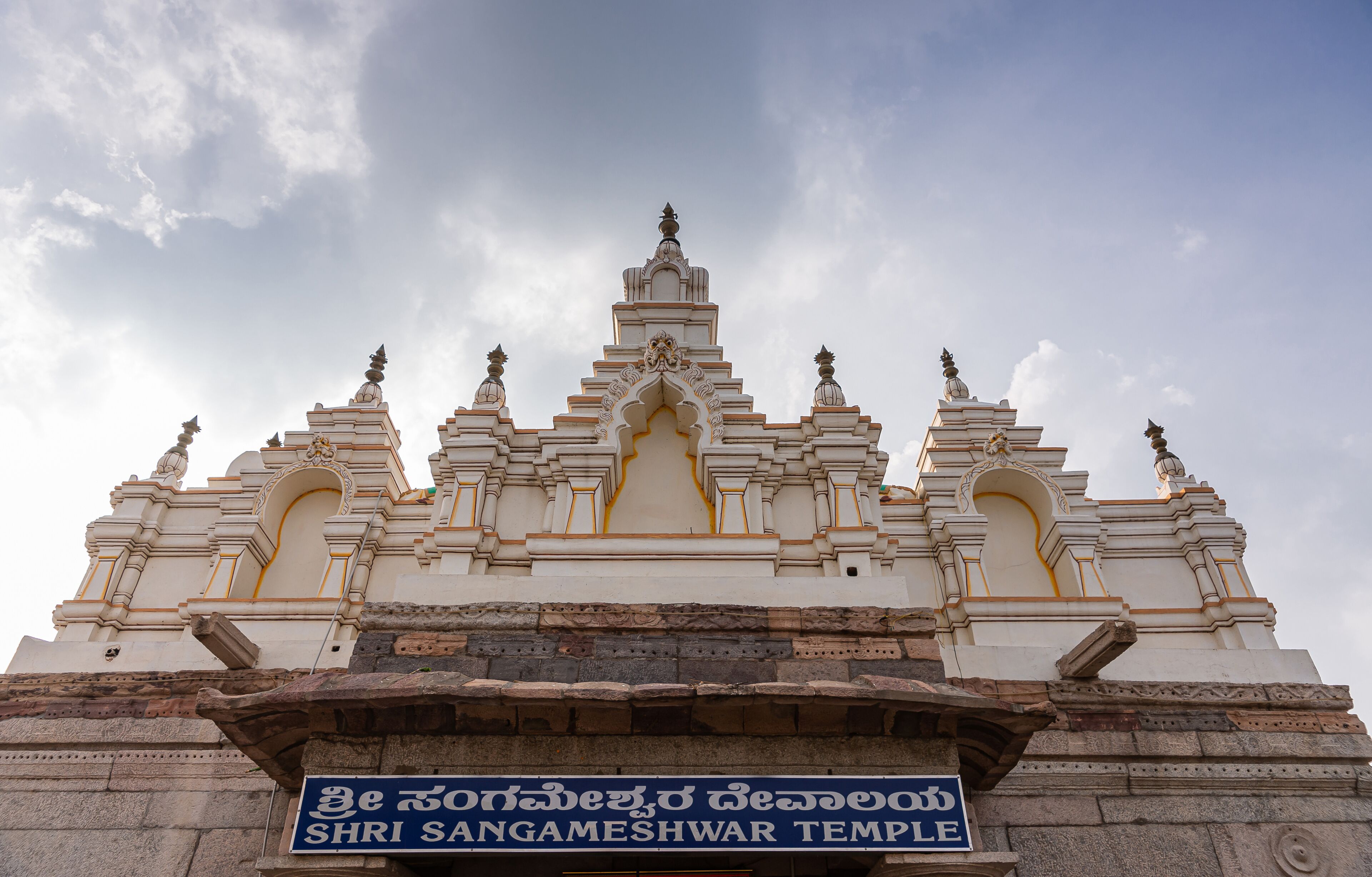 Bagalkot, Karnataka, India - November 8, 2013: Sri Sangameshwar Temple. Sculpted top in white with yellow trim on top of brown stone building under gray cloudscape.