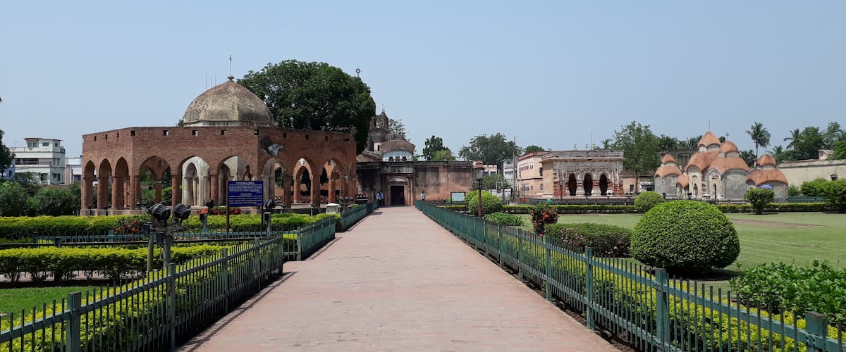 A pathway leading to a historic building with arches and domes, surrounded by well-maintained gardens and a clear sky.