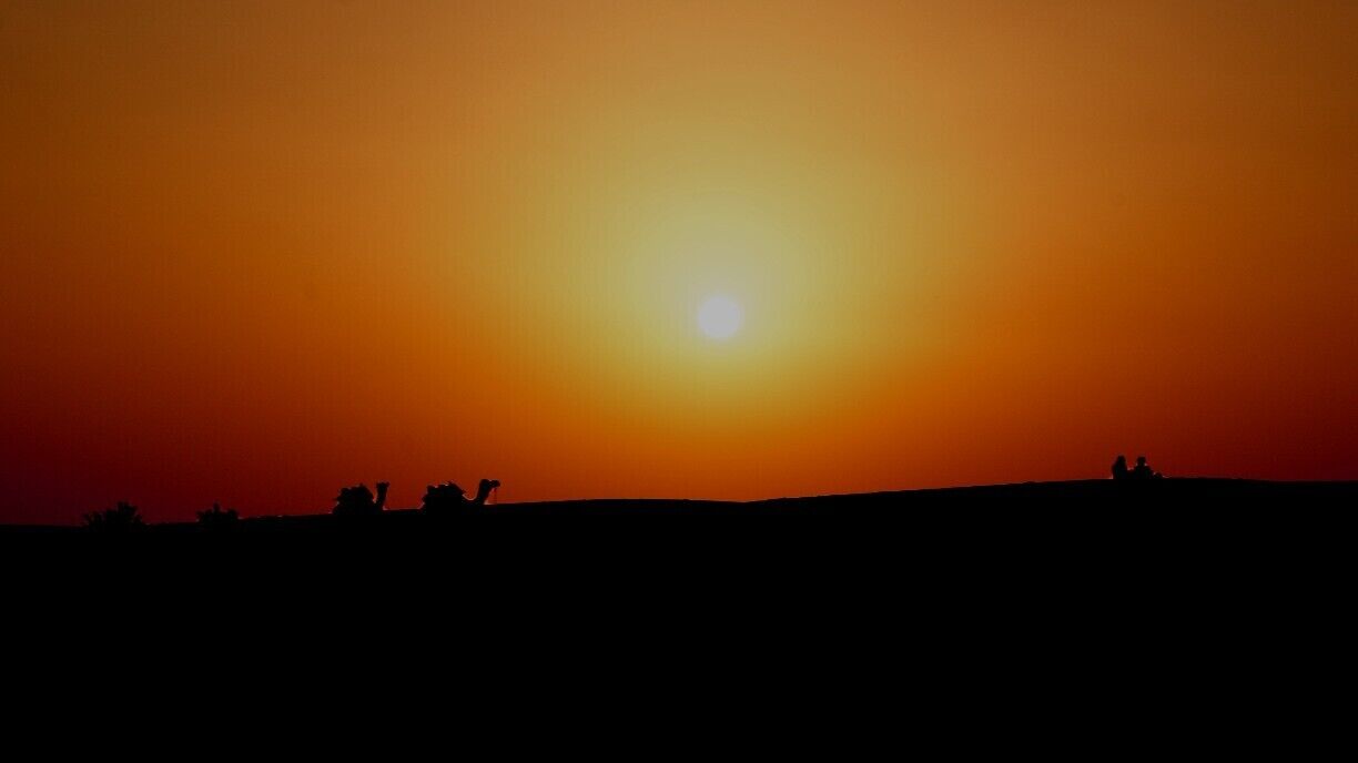 A sunset over a silhouette of a hill with people and camels.
