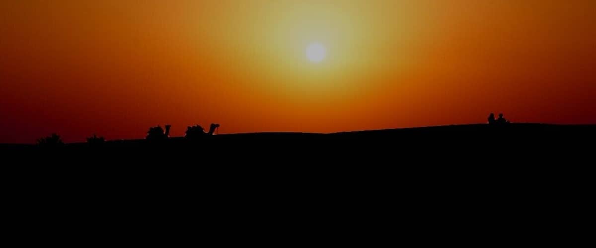 A sunset over a silhouette of a hill with people and camels.