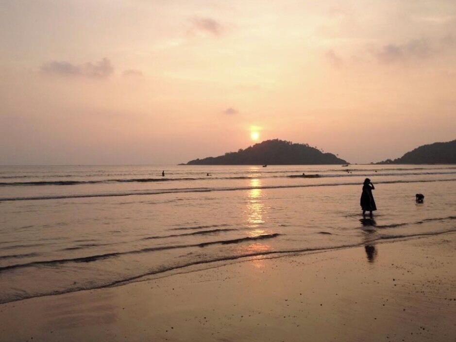 A beach at sunset with calm waters, a person walking, and a dog.