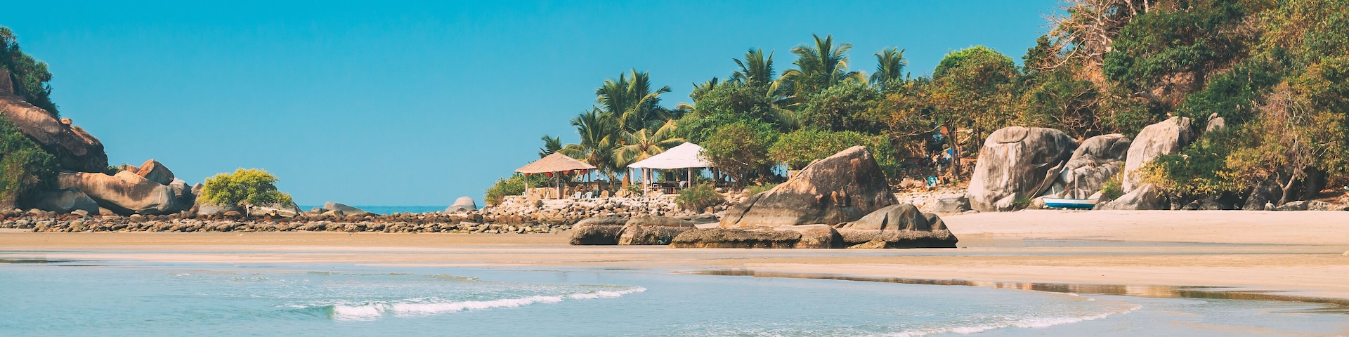A beach with clear blue water, rocky outcrops, and palm trees.