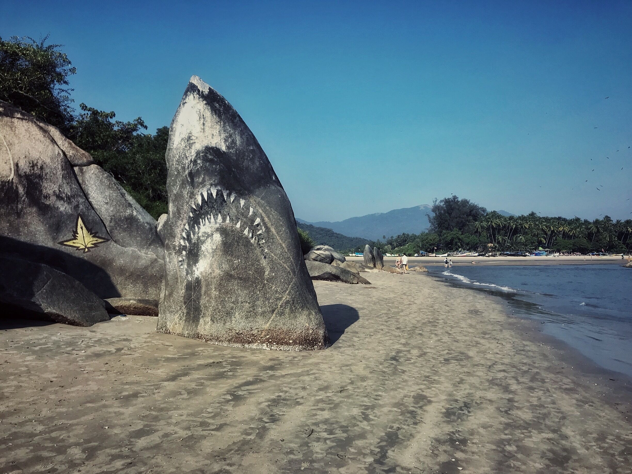 Una gran roca con un símbolo de hoja, una playa arenosa y palmeras al fondo.