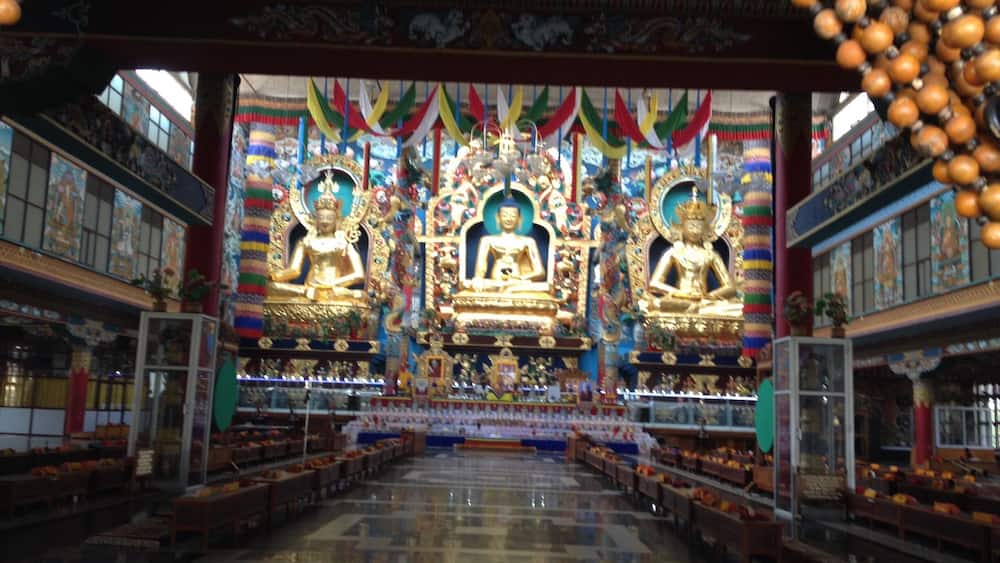 A temple interior with three golden statues, colorful drapes, and rows of benches.