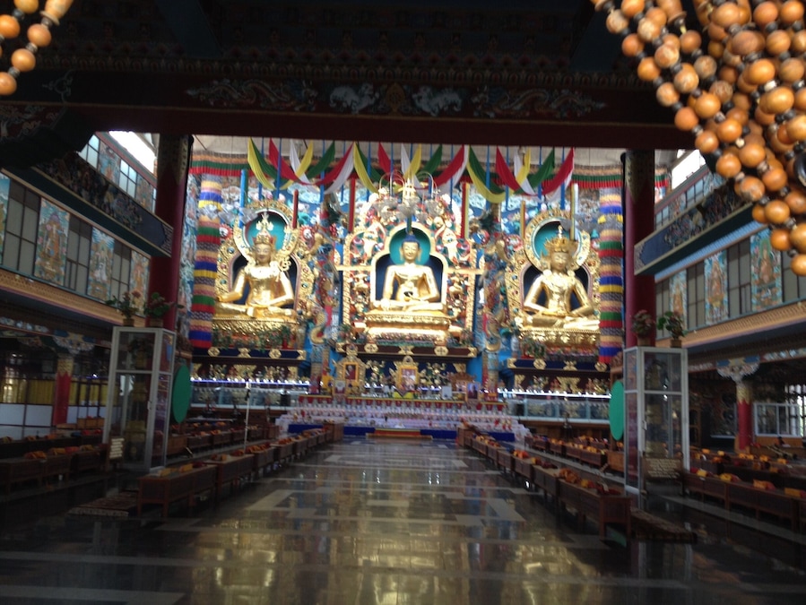 A temple interior with three golden statues, colorful drapes, and rows of benches.