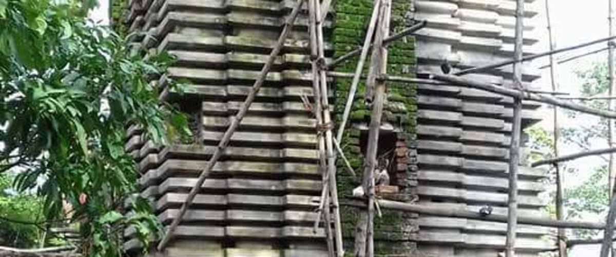A large, ancient stone structure with a staircase leading up to a doorway, surrounded by scaffolding and greenery.