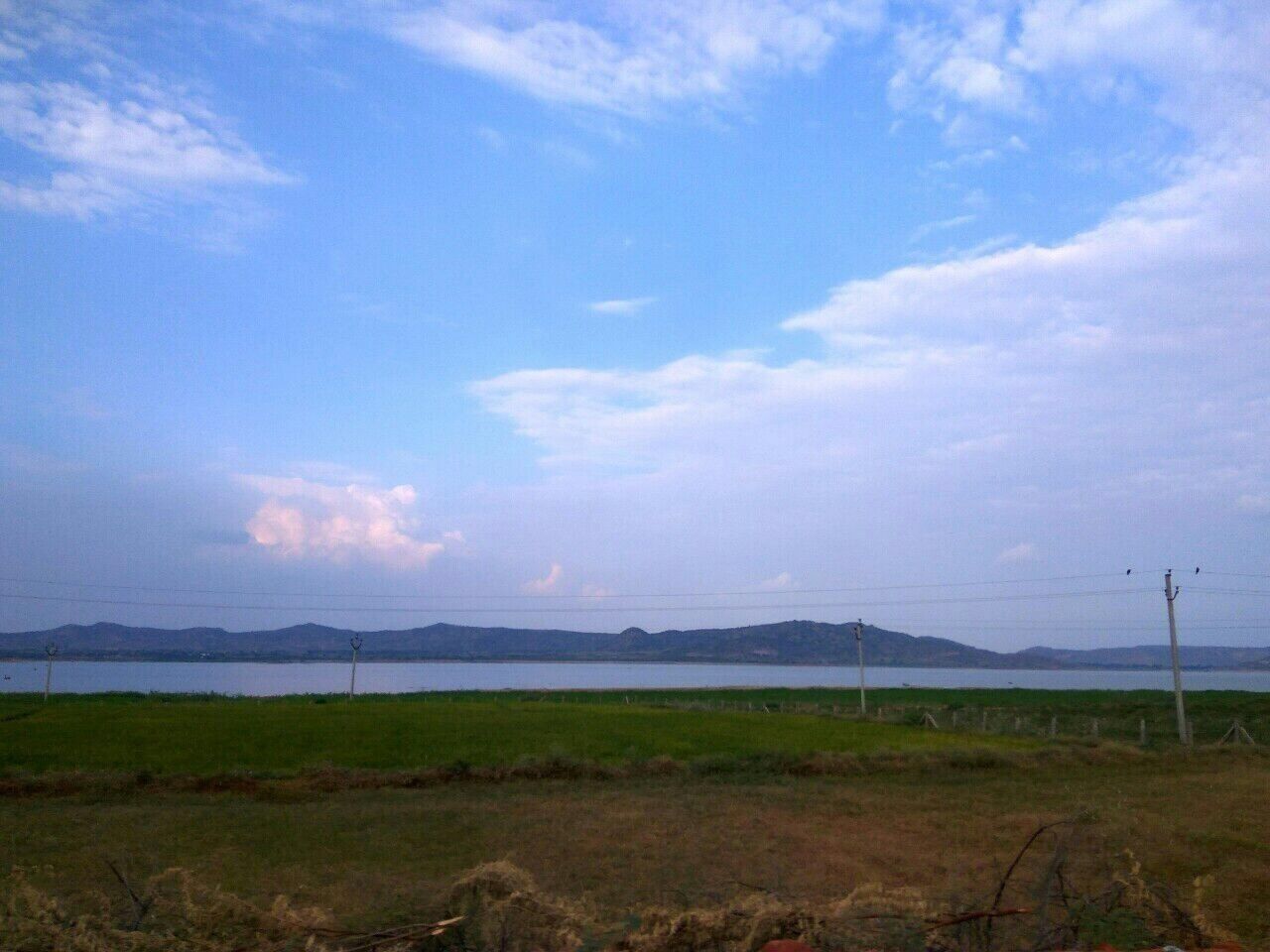 A lake with mountains in the background, surrounded by grassy fields and utility poles.