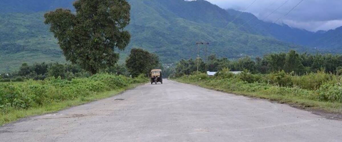 A rural road with a truck in the distance, surrounded by lush green hills and a cloudy sky.