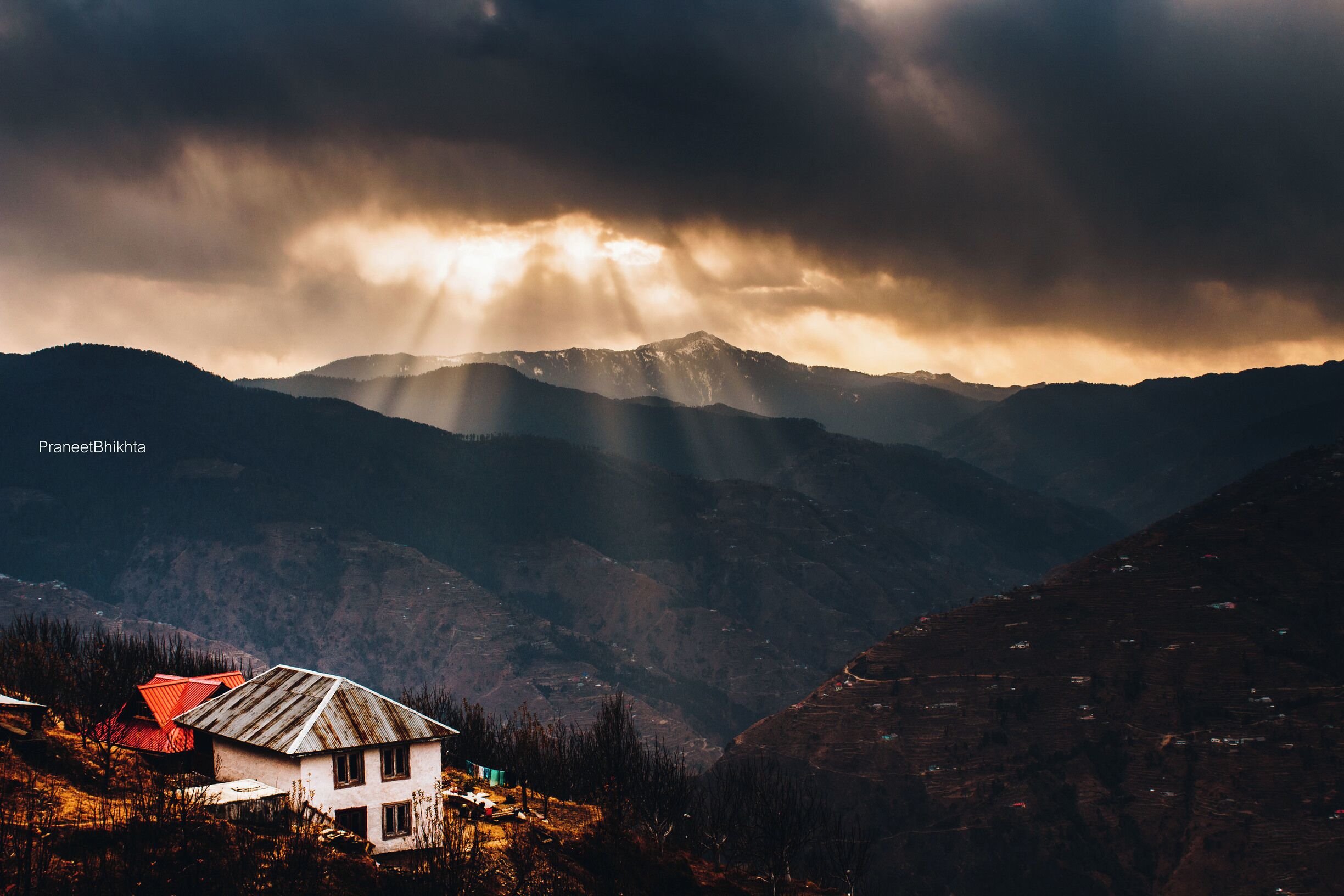 A mountainous landscape with a house in the foreground and rays of sunlight piercing through clouds.